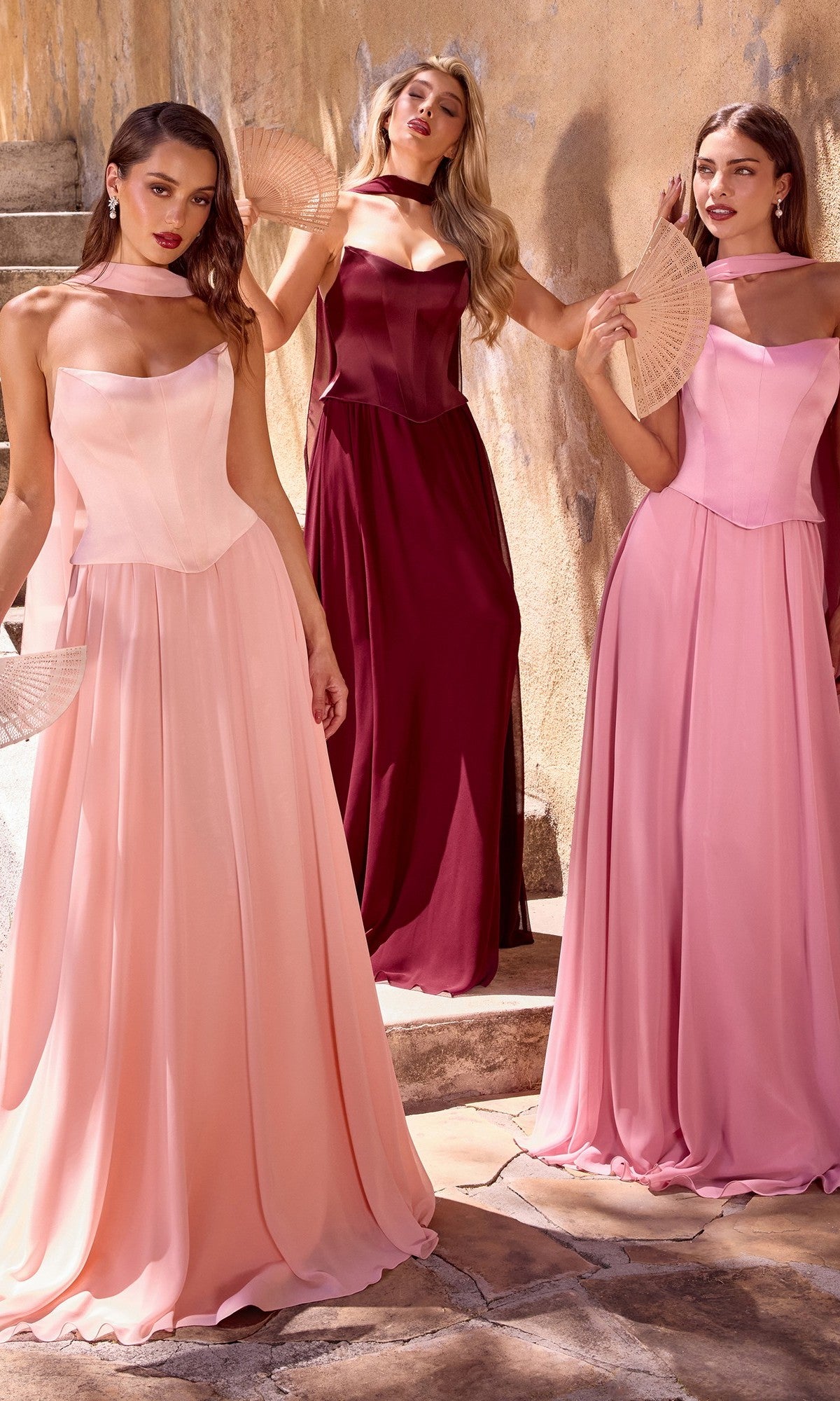Three women in floor-length dresses stand by a textured wall, holding fans. Two wear light pink formal dresses, while one shines in the Ladivine CD386 Strapless Long Corset Prom Dress in deep burgundy.